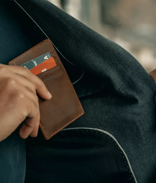 Close up of one hand holding a suit jacket open while the other slides a leather cardholder near the interior pocket; two bank cards stored in the slots.