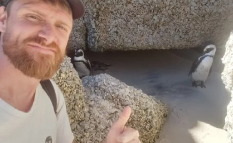 Student G. Behrends taking a selfie giving a thumbs up with two penguins among rocks stand behind.