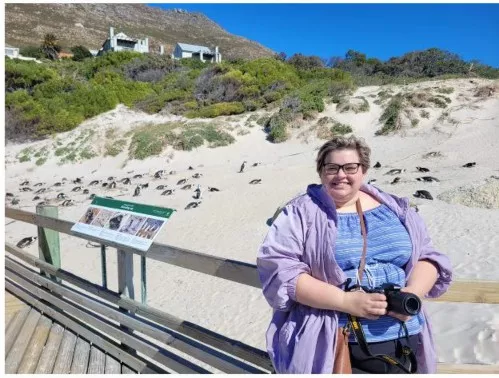 Joelle stands on a boardwalk with penguins lounging on the sand in the background.
