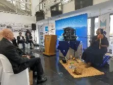 People sit across from each other and speak into microphones in a building. 