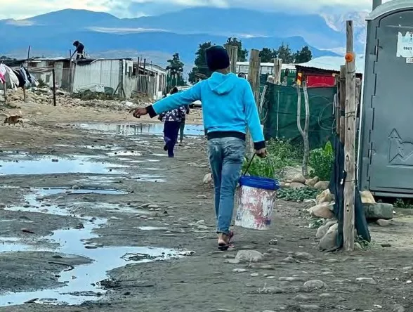 A person walks away from the photographer carrying a heavy bucket in an area of informal housing structures.