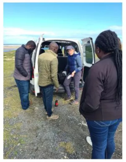 Four people stand behind the open back doors of a van on a grassy, rocky area