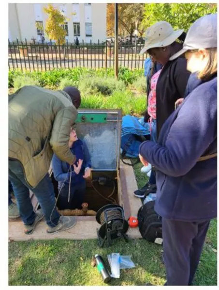 People gather around an access door in the ground while one person stands inside