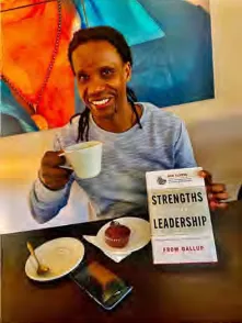 A person enjoys a warm beverage and snack while holding a book titled "Strengths Leadership"