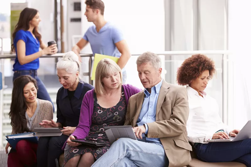 Crowd of people; man and woman conversing in background; foreground features man and woman of advanced age looking at a tablet, with the man pointing at the screen; pair of women seated to the side looking at a tablet together; woman seated other side typing on laptop.