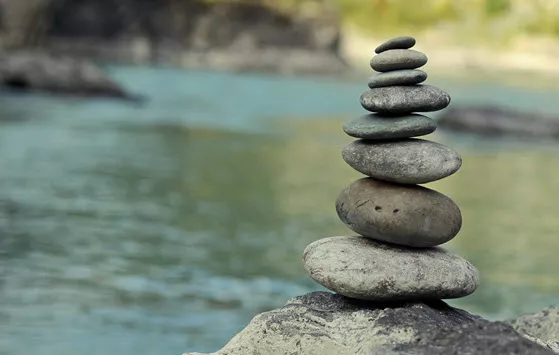 Cairn stack of pebbles atop a craggy rock in a body of water; shoreline, grassy hill out of focus in background
