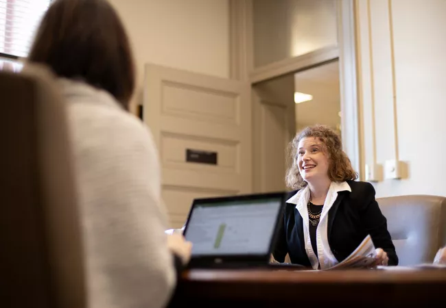 Two people sit across from each other at a conference room table, one with an open laptop, typing, while the other has paperwork in hand