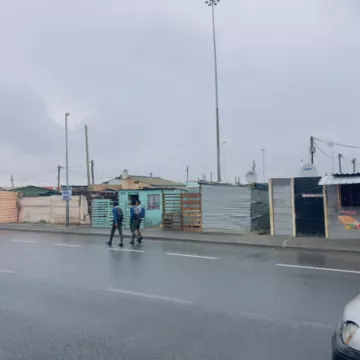 Two people walk on the street on a cloudy day, with small buildings in the background next to the road